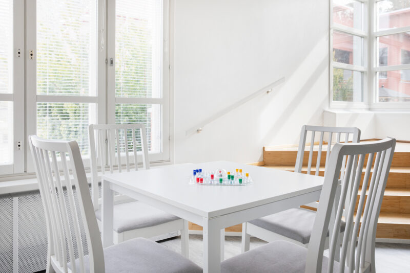 Valkoinen pöytä ja neljä tuolia sen ympärillä. Pöydällä on Kimble-peli. Pöydän takana avaria ikkunoita.