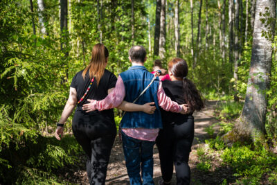 Neljä henkilöä kävelee metsässä polulla, kolme heistä pitää toisiaan olalta kiinni. Kaikkien selät ovat kameraan päin.