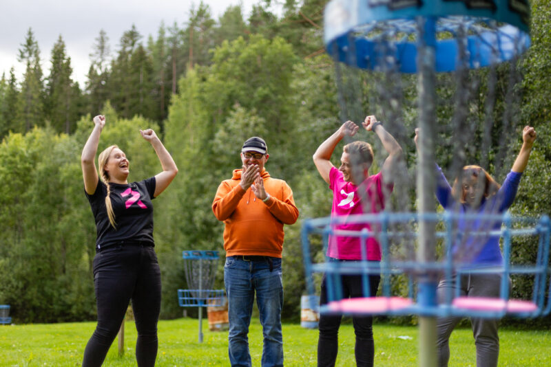 Henkilöt hurraavat käsiä ilmaan nostaen ulkona frisbeegolfmaalin äärellä. Taustalla näkyy vehreää metsää.